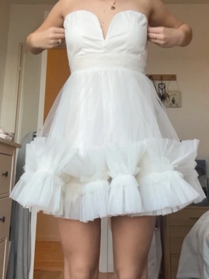 Vit klänning - Superfin vit kort klänning i tyll med hjärtformad överdel och markerad midja. Klänningen är axelbandslös och har stora dekorativa rosetter längst ner på kjolen för en extra söt look. Perfekt som student klänning! Klänningen är från ASOS! 