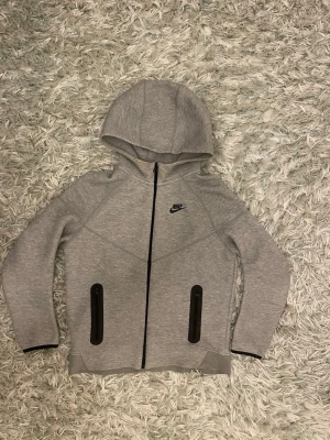 Grå Nike hoodie med dragkedja - Snygg grå hoodie från Nike med svart dragkedja framtill och två svarta dragkedjeförsedda fickor. Klassisk Nike-logga på bröstet och huva för extra stilpoäng. Tillverkad i mjukt och stretchigt material som känns skönt mot huden.