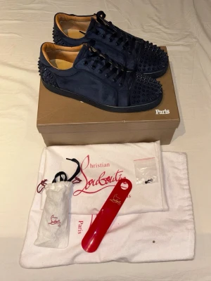 Mörkblå sneakers från Christian Louboutin - Snygga mörkblå sneakers från Christian Louboutin med coola svarta nitar på tå och häl. Skorna är gjorda i mocka och har klassiska snörningar. Insidan är ljusbrun. Har ett litet hål som syns på bild 5. Priset är inte fast skulle kunna tänka mig att gå ner. Perfekt för dig som vill sticka ut med exklusiva detaljer.