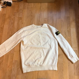 Vit sweatshirt från Stone Island - Vit sweatshirt från Stone Island med broderad logotyp på bröstet och klassisk svart logotyp-patch på vänster ärm. Tröjan har rund halsringning, ribbade muddar och är tillverkad i mjuk bomull. Perfekt för en stilren och avslappnad look.