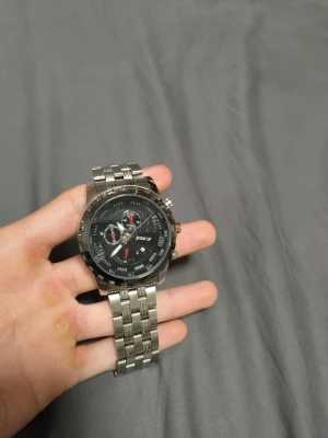 Silverfärgad klocka från Positif - Säljer en snygg och robust klocka från Positif med silverfärgat metallarmband och svart urtavla. Klockan har flera små urtavlor och röda detaljer, vilket ger den ett coolt och sportigt utseende. Perfekt för dig som gillar stilrena accessoarer.