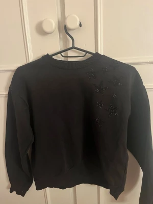 Svart sweatshirt med stjärndetaljer - Cool svart sweatshirt med rund hals och långa ärmar. På ena axeln finns flera svarta stjärnor i pärlor som ger en unik touch. Tröjan har ribbade muddar vid ärmslut och nederkant. Perfekt för dig som gillar enkla men snygga detaljer.