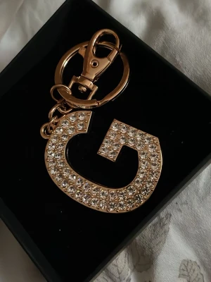Guldig nyckelring med strass G - Snygg nyckelring i form av bokstaven G, täckt med glittrande strass och guldfärgad metall. Perfekt att hänga på väskan eller nyckelknippan för en extra touch av bling.