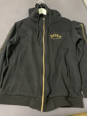 Svart hoodie från Hugo Boss - Snygg svart hoodie från Hugo Boss med dragkedja i guld och guldfärgade detaljer. Märket syns tydligt på bröstet och ärmarna har diskreta guldfärgade ränder. Huva med dragsko och två fickor framtill. Perfekt för en stilren och avslappnad look.