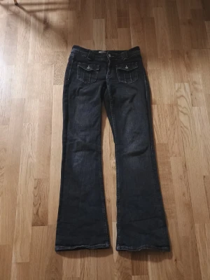 Svart bootcut jeans med fickor - Snygga svarta bootcut jeans med två framfickor med lock och knappar samt klassiska bakfickor. Jeansen har en låg midja och utsvängda ben. Perfekta för en avslappnad och trendig look.