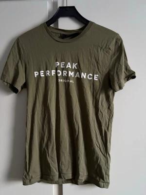 Grön Peak Performance t-shirt - Snygg grön t-shirt från Peak Performance med vit logga tryckt på bröstet. Klassisk rund halsringning och korta ärmar. Endast använd 1 gång men skrynklig.