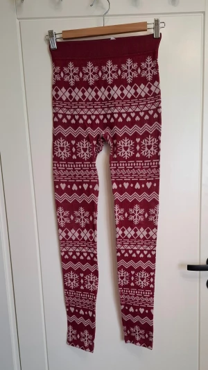 Vinröda leggings med snöflingor från Rusta  - Supersköna vinröda leggings med vitt vintermönster och snöflingor från Rusta. Tillverkade i 96% polyester och 4% elastan för en stretchig och bekväm känsla. Hög midja och åtsittande passform, perfekta för mysiga hemmakvällar. 