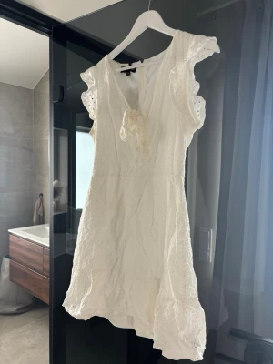 Vit kort klänning med volangärm - Söt vit kortklänning med volangärmar och broderade detaljer. Klänningen har v-ringning med knyt framtill och är tillverkad i ett lätt bomullstyg. 