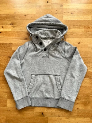 Vintage Abercombrie And Fitch/Hollister hoodie med knappar - Säljer nu denna snygga gråa vintage Hoodien från Abercombrie and Fitch. Tröjan är i väldigt fint skick och ser i princip helt ny ut. Modellen på bilden är 183 och väger 67 kilo. Tröjan är i storleken M. Skriv gärna vid mer funderingar:)