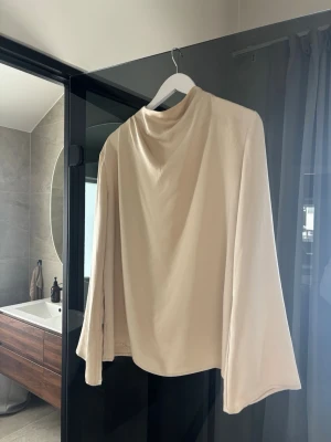 Beige långärmad topp med lös passform - Stilren beige topp med lång ärm och lös passform. Toppen har en mjuk, slät yta och en enkel design utan synliga detaljer. 