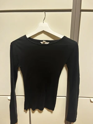 Svart långärmad topp i stretch - En enkel och stilren svart långärmad topp med rund halsringning. Tillverkad i ett tunt, stretchigt material som sitter luftigt mot kroppen. Perfekt att styla med jeans eller kjol för en clean look.