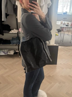 Svart axelväska med kedjedetaljer - Cool svart axelväska med glansigt material och silverfärgade kedjor längs kanterna och som axelband. Väskan har en mjuk och slouchy form, perfekt för att bära över axeln. 