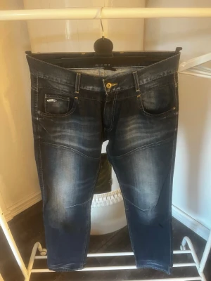 Mörkblå jeans med slitningar - Säljer ett par mörkblå jeans med snygga slitningar och kontrastsömmar. Jeansen har fem fickor, guldfärgad knapp och coola detaljer på bakfickorna. Passar perfekt till en avslappnad streetstil. W33 L32. Hör av er vid frågor!