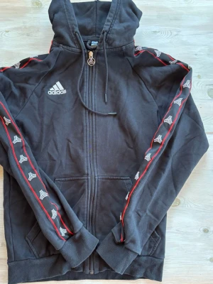 Svart Adidas hoodie med dragkedja - Svart hoodie från Adidas med dragkedja framtill och vit logga på bröstet. Röd och vit detalj längs ärmarna och fickor på sidorna. Huva med dragsko och ribbade muddar. Perfekt för en sportig och avslappnad stil.