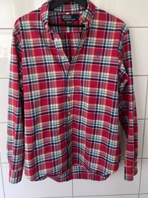Rutig röd skjorta från Polo Ralph Lauren - Klassisk rutig skjorta från Polo Ralph Lauren i rött, blått och vitt. Skjortan har lång ärm, knappar framtill och button-down krage. Tillverkad i mjuk bomull och har den ikoniska loggan broderad på bröstet.