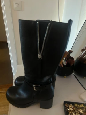 Svarta chunky stövlar med spänne - Snygga svarta stövlar med grov sula och klack. Stövlarna har ett dekorativt spänne vid ankeln och dragkedja på insidan. Tillverkade i ett läderliknande material med högt skaft och rundad tå. Perfekta för en edgy look.