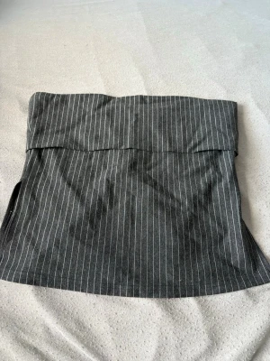 Mörkgrå randig tubtopp med slitsar  - Säljer en mörkgrå bandeau tubtopp med tunna vita ränder. Toppen är ärmlös och har en enkel, stilren design med slits i sidan. Toppen har en croppad passform.