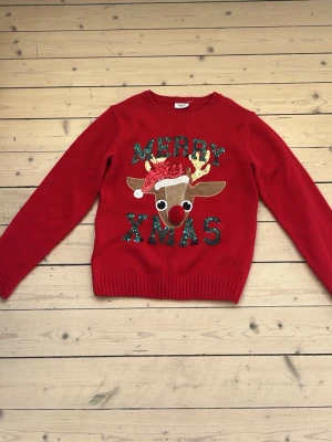 Röd jultröja med renmotiv - Rolig röd stickad jultröja med texten 'Merry Xmas' i grönt glitter och ett stort renmotiv med 3D-detaljer som röd nos och tomteluva. Perfekt för att sprida julstämning! Långa ärmar och ribbade muddar.