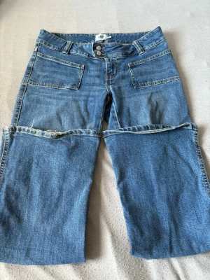 Blå lågmidjade jeans - Fina blå jeans från  Lager 157. Raka ben, dubbla knappar i midjan och stora fickor fram och bak. Tillverkade i klassisk denim med cool vintage vibe, de enda skadorna är de som är längst ner på benet som bilden visar. Längden är ”short”