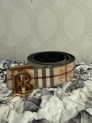 Beige rutigt bälte med guldspänne 105cm - Snyggt bälte i beige med klassiskt rutigt mönster och ett stort guldigt B-spänne. Perfekt accessoar för att lyfta din outfit med en trendig touch. Bältet har en exklusiv känsla och passar till både jeans och byxor.