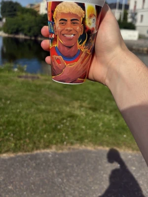 Färgglad plastmugg med fotbollsmotiv - Säljer en färgglad plastmugg med tryck av en fotbollsspelare i röd tröja och blå shorts, omgiven av eld och stadionmiljö. Muggen har vita kanter och livfulla illustrationer runt om hela ytan. Perfekt för dryck på picknick eller hemma.