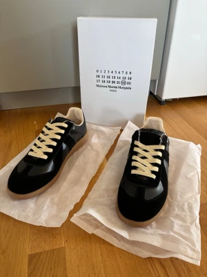 Maison Margiela svarta sneakers - Svarta sneakers från Maison Martin Margiela med beigea skosnören och gummisula. Skorna har en klassisk låg siluett, ovandel i mocka och läder samt diskreta detaljer. Perfekta för dig som gillar stilrena och tidlösa sneakers.