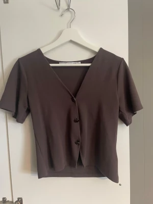 Brun kortärmad topp med knappar - Snygg brun topp med v-ringning och tre knappar framtill. Modellen är kortärmad och har en enkel, stilren design som passar till mycket. Materialet känns mjukt och stretchigt. Perfekt för dig som gillar minimalistisk stil.