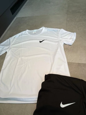 Nike träningsset t-shirt och shorts - Säljer ett fräscht träningsset från Nike med en vit t-shirt och svarta shorts. Båda plaggen har den klassiska Nike-loggan framtill. Materialet är lätt och andas, perfekt för träning eller sportaktiviteter.