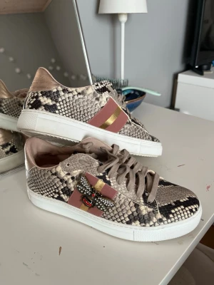 Sneakers med ormmönster - stokton  - Snygga sneakers med beige och svart ormmönster, breda ränder i rosa och guld på sidan samt en cool bi-applikation med stenar. Skorna har beige snörning och vit sula. I märket stokton 