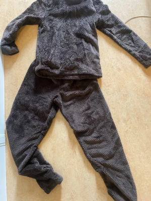 Mörkbrunt mjukisset loungewear - Mysigt tvådelat loungewear-set i mörkbrun färg med långärmad tröja och matchande byxor. Setet är gjort i ett mjukt, strukturerat tyg som känns skönt mot huden. Perfekt för att chilla hemma i och hålla sig varm under kalla dagar.