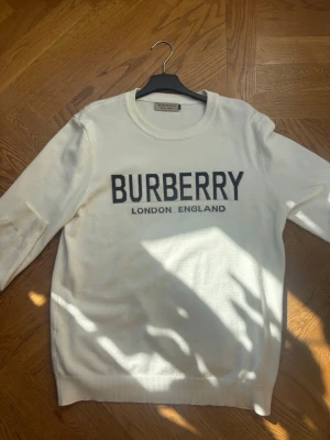 Vit stickad tröja från Burberry - Snygg vit stickad tröja från Burberry med svart text 'BURBERRY LONDON ENGLAND' på bröstet. Tröjan har rund halsringning och långa ärmar. Perfekt för dig som gillar klassisk och stilren design. (Står XL men sitter som en M)