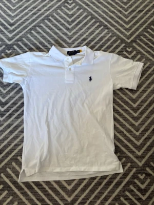 Vit pikétröja från Ralph Lauren - Klassisk vit pikétröja från Ralph Lauren med korta ärmar, krage och knappar framtill. Tröjan har en blå broderad logga på bröstet och är gjord i mjuk bomull för en clean och tidlös look. Säljer pågrund av att den inte kommer till användning och jag aldrig använt den och den bara hängt i garderoben därav ett superpris 449kr!!! Nypris 1700 kr 