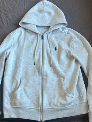 Ljusgrå hoodie med dragkedja Polo Ralph Lauren  - Ljusgrå hoodie med dragkedja från Polo Ralph Lauren, logga på bröstet. Mjuk bomullsblandning som är skön att ha på sig och passar perfekt till en avslappnad stil. Personen är ca 180cm men är lite liten så passar dig perfekt som är lite under.