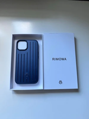 Blått mobilskal från Rimowa iphone 13/14 - Snyggt blått mobilskal från Rimowa med stilrena vertikala ränder och diskret logga längst ner. Skalet är gjort i ett tåligt material och har en modern, minimalistisk design som ger din mobil en exklusiv känsla.