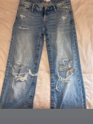 Ljusa ripped jeans från Gina Tricot - Säljer ett par ljusblå jeans från Gina Tricot med stora slitningar och hål på båda benen. Modellen är rak och har klassiska fem fickor samt bälteshällor. Perfekta för en avslappnad och trendig look.