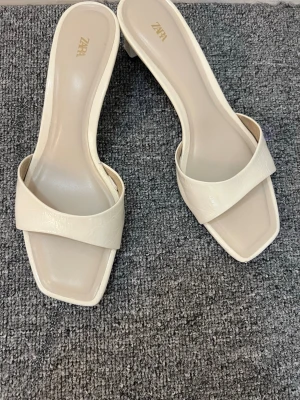 Krämvita slip-in pumps från Zara - Stilrena krämvita slip-in pumps från Zara med fyrkantig tå och låg klack. Skorna har bred rem över foten och är tillverkade i ett glansigt, konstläderliknande material. Perfekta för dig som gillar minimalistisk och trendig design.