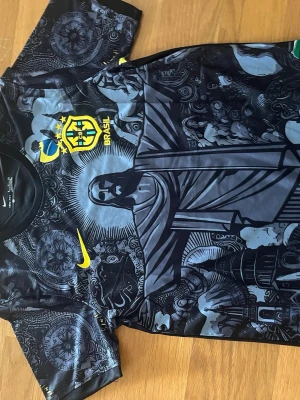 Nike Brasil svart tröja  - Snygg svart tröja  från Nike med Brasil-logga och gult Nike-märke. Framsidan har ett detaljerat grafiskt tryck med jesus  och brasilianska symboler. Korta ärmar med mönster och flagga på axlarna. Perfekt för fotboll eller streetwear.