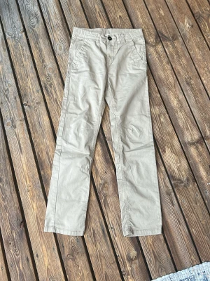 Beiga chinos från Kappahl  - Snygga beiga chinos från Kappahl med raka ben och klassisk passform. Byxorna har två framfickor och en enkel knappstängning i midjan. Perfekta för en avslappnad och stilren look. Materialet känns mjukt och bekvämt.