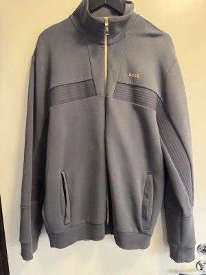 Grå zip hoodie från BOSS - Snygg grå hoodie från BOSS med dragkedja framtill och hög krage. Hoodien har diskreta ribbade detaljer över bröstet och två fickor på sidorna. Minimalistisk design med BOSS-logga i vitt på bröstet. Perfekt för en clean och stilren look. Står XXL men passar XL