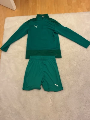 Grönt träningsset från Puma - Snyggt grönt träningsset från Puma med långärmad tröja och matchande shorts. Båda plaggen har Puma-loggan tryckt i vitt. Materialet är lätt och andas, perfekt för träning eller fotboll. Tröjan har hög krage och dragkedja vid halsen. Pris kan diskuteras vid snabb affär!