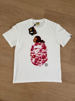 Vit BAPE t-shirt med apa och camomönster - Vit t-shirt från BAPE med klassisk apa-tryck ovanför ett rosa och rött camomönstrat ansikte på bröstet. T-shirten har rund hals och korta ärmar. Materialet är mjuk bomull och etiketten visar BAPE-loggan.