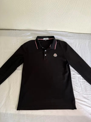 Svart långärmad piké från Moncler - Snygg svart långärmad pikétröja från Moncler med klassisk krage och knäppning framtill. Tröjan har Moncler-logga på bröstet och detaljer i rött, vitt och blått på krage och ärmslut. Perfekt för en stilren och sportig look.