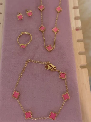 Smyckeset med rosa fyrklöver - Säljer ett smyckeset i rostfritt stål med halsband, armband, ring och örhängen. Alla delar har guldfärgad kedja och dekoreras med rosa fyrklöverformade hängen. Perfekt för dig som gillar färgglada accessoarer och vill sticka ut lite extra.