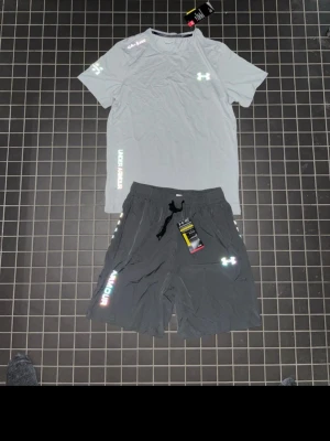 Under Armour träningsset grå/svart - Snyggt träningsset från Under Armour med en ljusgrå t-shirt och svarta shorts. T-shirten har logga på bröstet och tryck på ärmen, medan shortsen har dragsko i midjan, sidofickor och logga samt text längs benet. Perfekt för gymmet eller löpning.