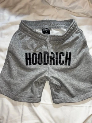 Grå shorts från Hoodrich med snörning - Grå mjukisshorts från Hoodrich med svart broderad logga framtill. Shortsen har elastisk midja med snörning och är tillverkade i mjukt bomullsmaterial. Perfekta för chill eller träning.