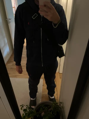 Mörkblå tracksuit från ralph lauren - Säljer en mörkblå tracksuit med dragsko i midjan och resår vid bensluten. Byxorna har en avslappnad passform och en liten röd logga på låret. Perfekta för chill dagar eller när du vill vara bekväm.