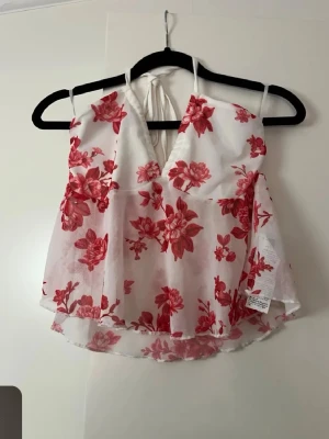 Vit och röd blommig linnetopp - Söt vit linnetopp med röda blommor och v-ringning. Toppen har tunna axelband och knyt i nacken för en luftig känsla. Perfekt för varma dagar och ger en romantisk vibe till din outfit.
