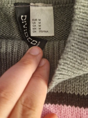 Grå stickad tröja med rosa ränder - Snygg stickad tröja i grått med rosa och svarta horisontella ränder. Tröjan har långa ärmar och ribbade muddar vid ärmslut och nederkant. Perfekt för dig som gillar färgglada detaljer i vardagslooken.