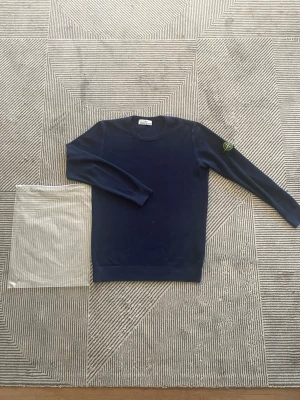Mörkblå stickad tröja Stone Island - Snygg mörkblå stickad tröja från Stone Island med rund hals och lång ärm. Tröjan har en diskret struktur i materialet och den klassiska Stone Island-loggan på vänster ärm. Perfekt till jeans eller chinos för en clean look.