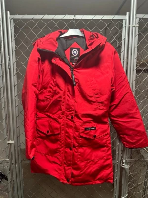 Röd parkas från Canada Goose - Snygg röd parkas från Canada Goose med huva och flera praktiska fickor framtill. Jackan har en klassisk längre modell och patch med logga på ärmen. Perfekt för kalla dagar och har en robust dragkedja samt tryckknappar. Pälsen på luvan saknas tyvärr.